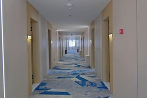 Um corredor de hotel comprido e estreito com carpete estampado, portas em ambos os lados e um aparelho de iluminação no teto.