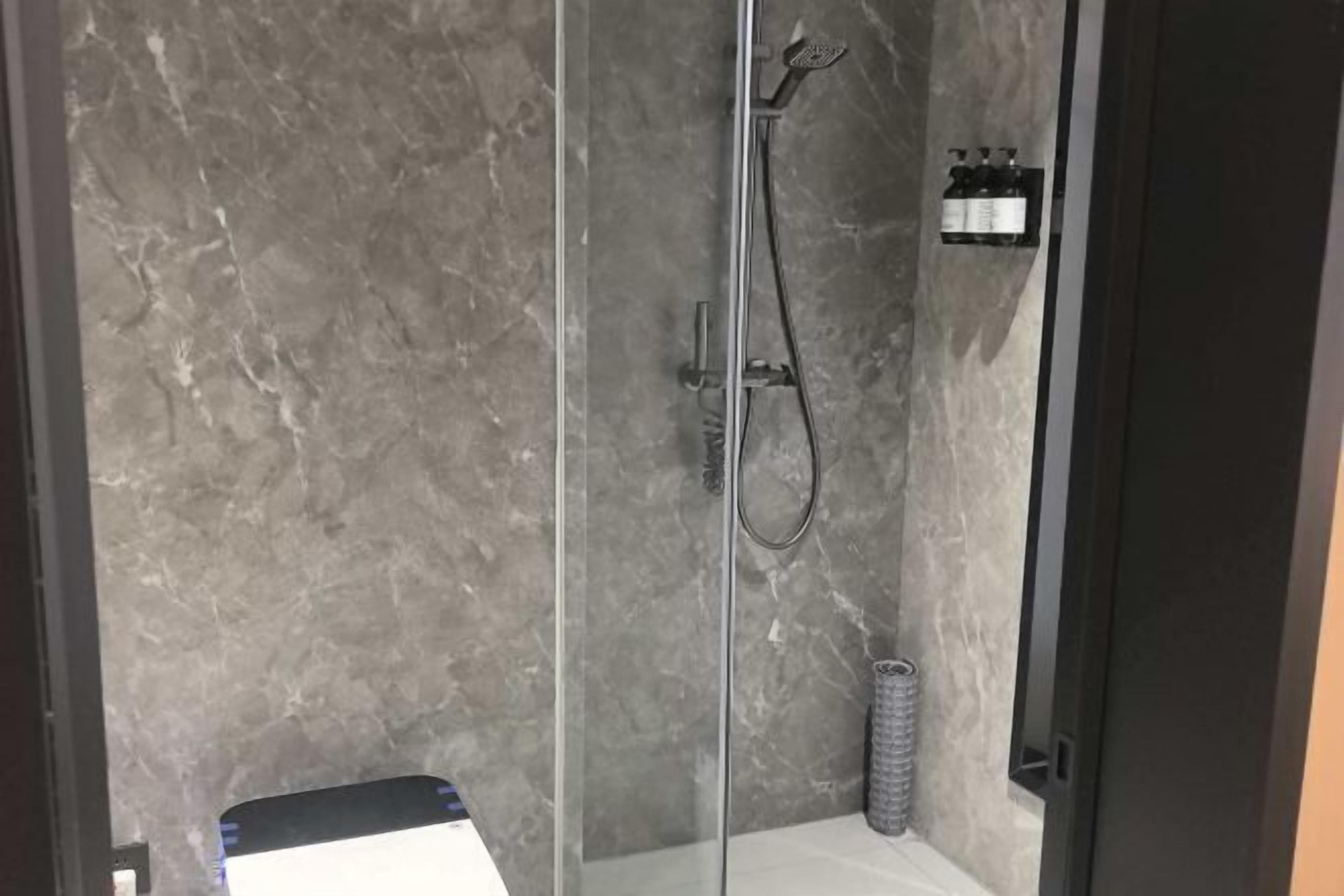 A shower area with a glass door, a handheld showerhead, and a bottle shelf.
