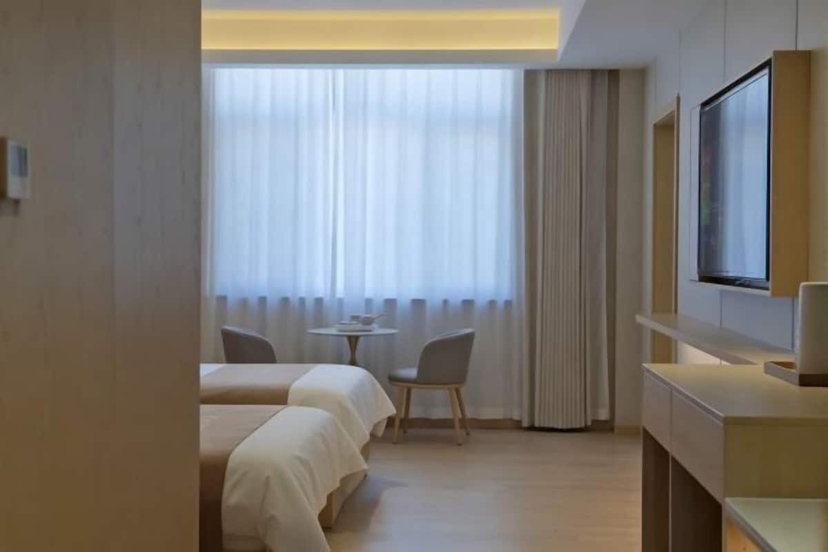 Habitación de hotel con dos camas, una mesita pequeña, una silla, televisor y un ventanal grande con cortinas.