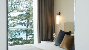 Habitación de hotel con cama, mesita de noche, lámpara y vista a árboles a través de una ventana.