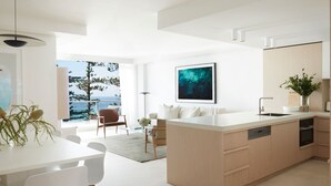 Una cocina moderna con una isla central, un comedor con mesa y sillas blancas, y una sala de estar con sofá y vista al mar.