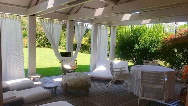 A covered outdoor seating area with wooden flooring, cushioned seating, and a table set for dining. - Holiday home mit Garten (Rosolina)