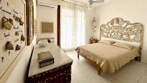 A bedroom with a bed, a ceiling fan, a nightstand, a decorative headboard, a wall-mounted air conditioner, and a wall with various items. - Holiday home mit Garten (Rosolina)
