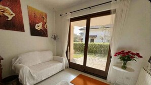 A room with a sofa, a small table with a vase of flowers, and a view of the outside. - Holiday home mit Garten (Rosolina)