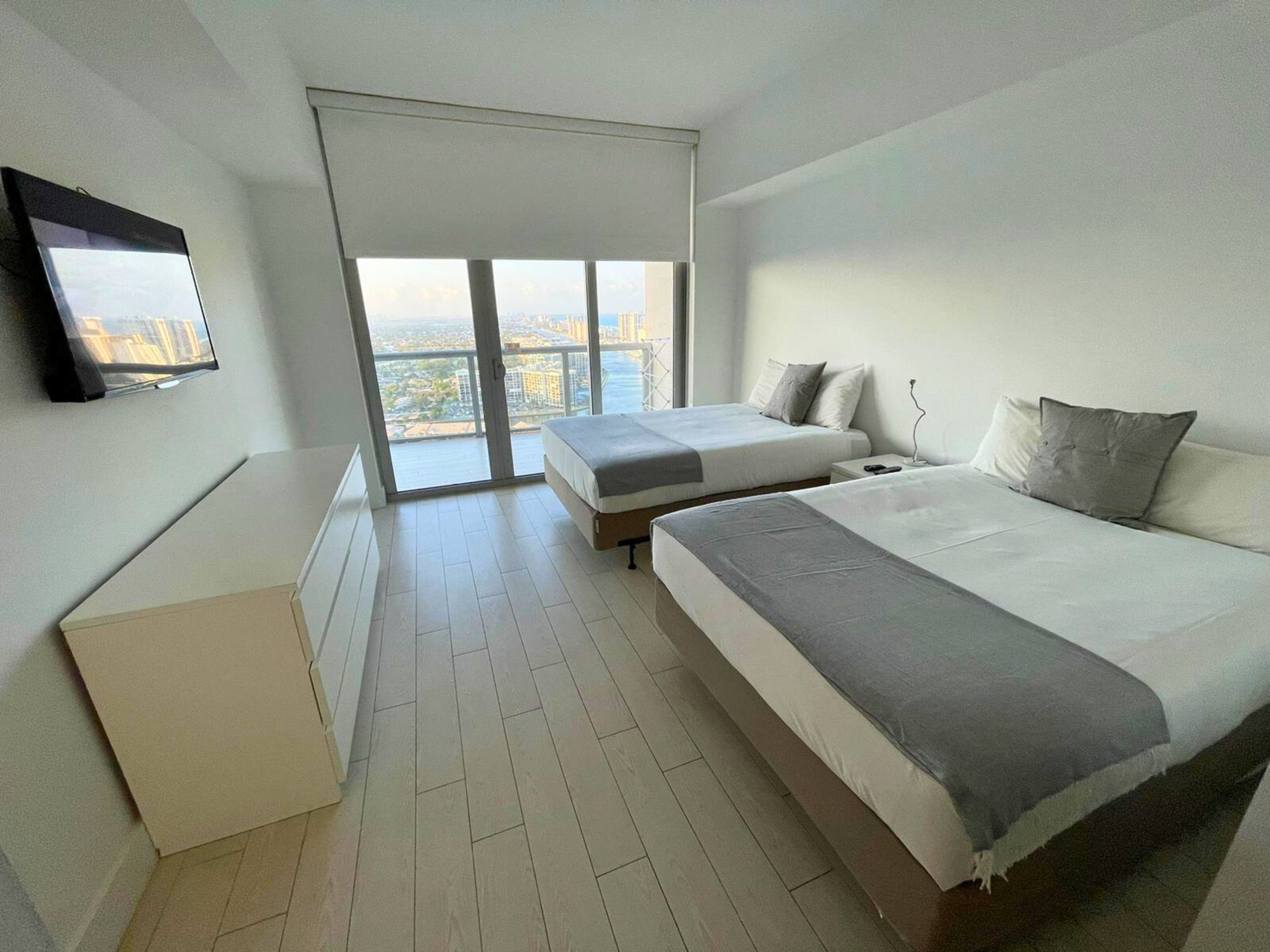 A modern bedroom with two beds, a TV, and a balcony with a city view.