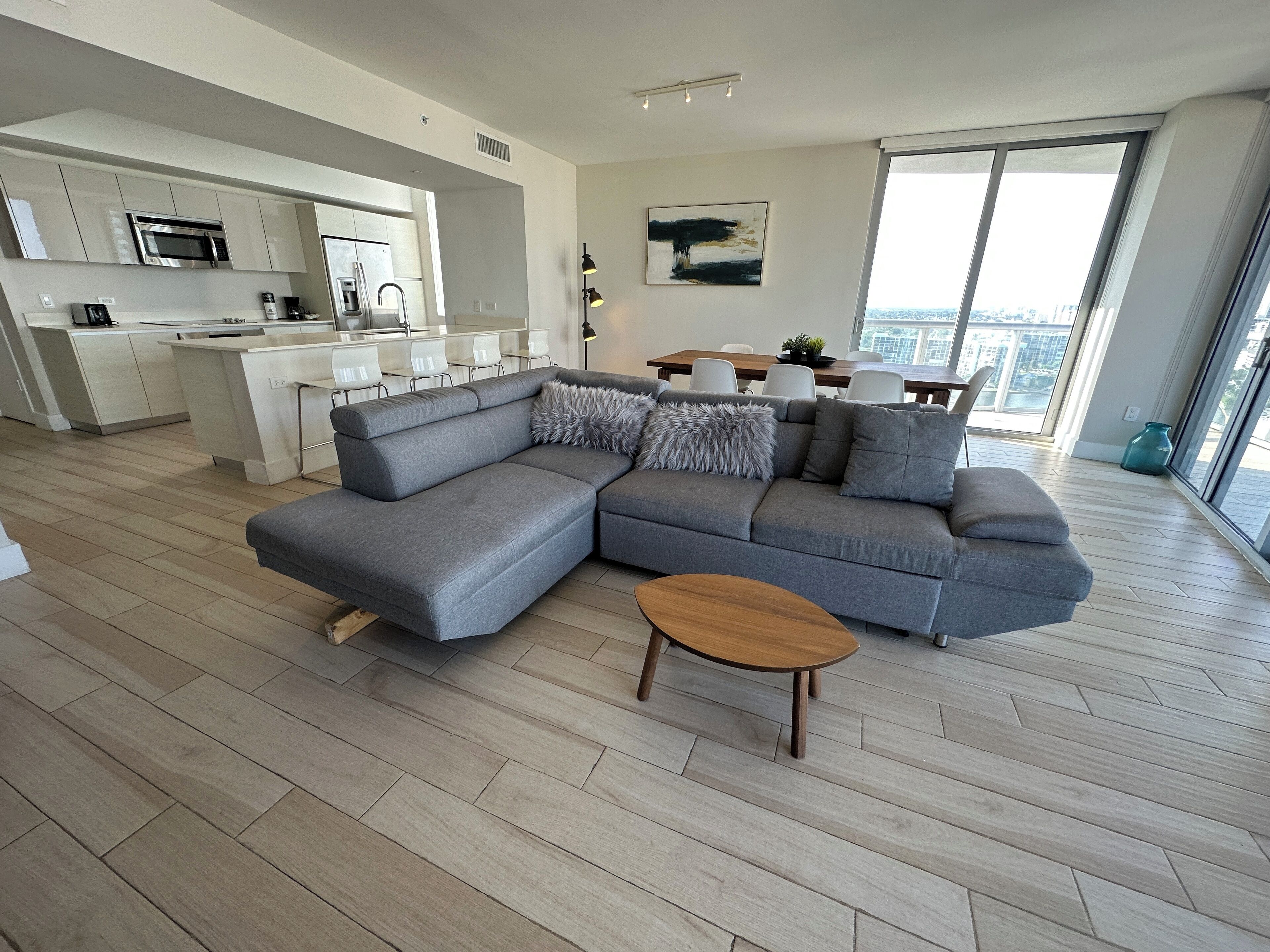 Una sala de estar moderna con un sofá seccional gris, una mesa de centro de madera y un comedor con un ventanal que ofrece vistas a la ciudad.