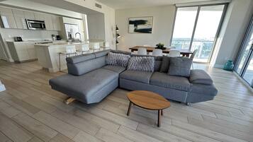 Una sala de estar moderna con un sofá seccional gris, una mesa de centro de madera y un comedor con un ventanal que ofrece vistas a la ciudad.