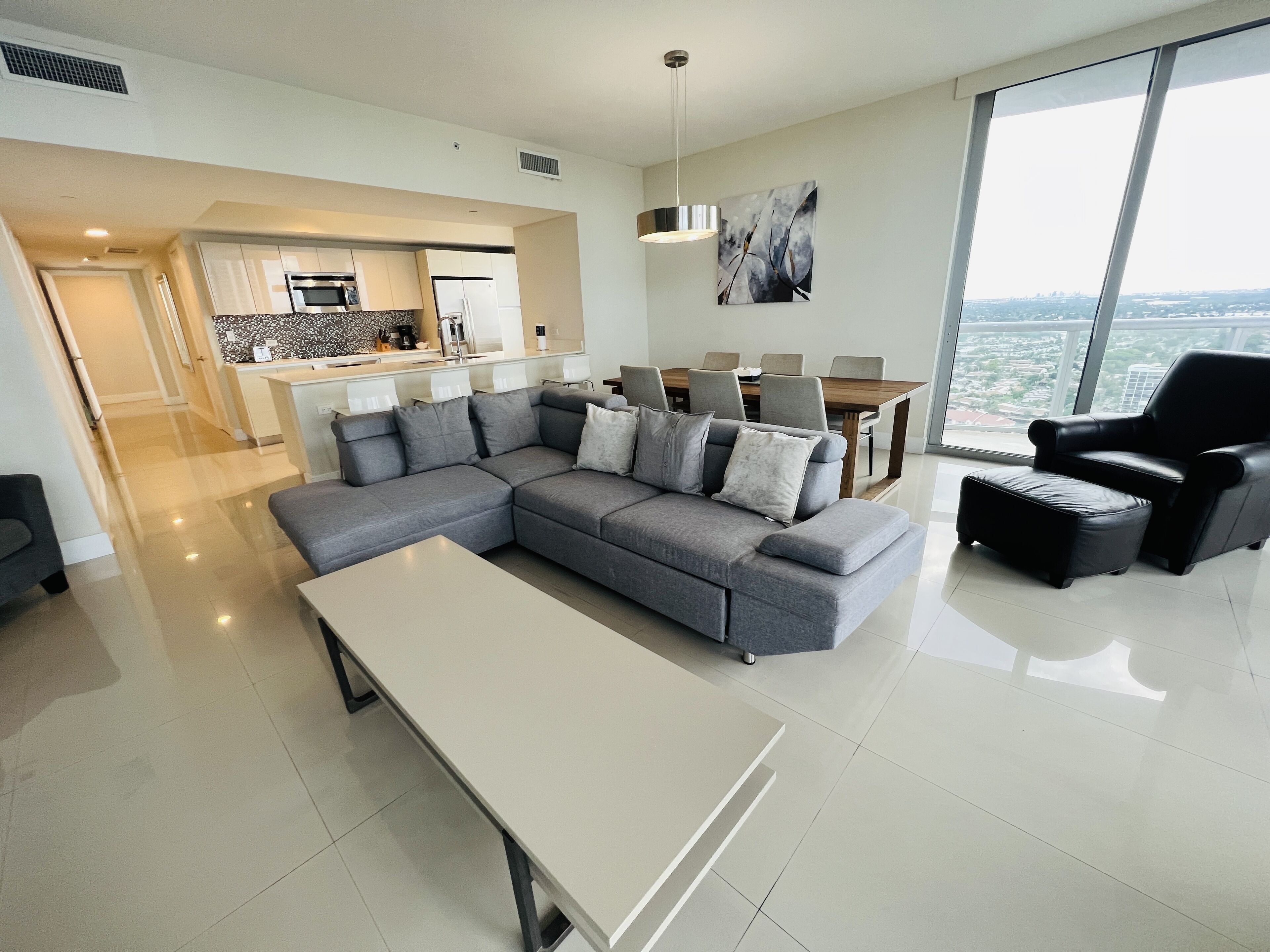 A modern living room with a grey sectional sofa, a white coffee table, and a black leather armchair. There is a dining area with a wooden table and chairs, and a kitchen in the background.