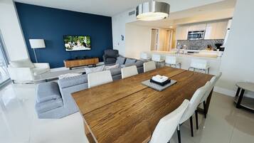 A modern living room with a wooden dining table, grey sofas, and a TV mounted on a blue wall.