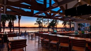 Un área de comedor al aire libre con mesas y sillas de madera, con vista a un cuerpo de agua al atardecer.
