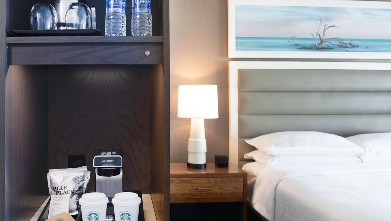 A hotel room with a bed, a nightstand with a lamp, a coffee machine, and a Starbucks coffee station.