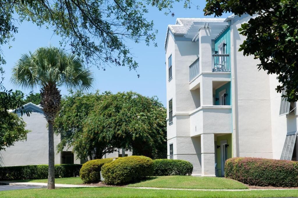 Un edificio de varios pisos con fachada blanca y detalles color turquesa, rodeado de jardines bien cuidados que incluyen una palmera y arbustos podados.