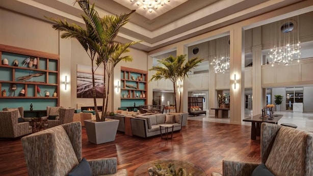 Eine geräumige Lobby mit hoher Decke, Holzböden und modernen Möbeln. Es gibt mehrere Sitzbereiche mit Sofas und Sesseln. Die Lobby verfügt über große Fenster und eine dekorative Deckenleuchte.