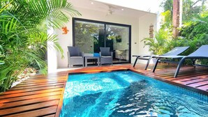 Una piscina con cubierta de madera, dos sillones de descanso y un ventanal de vidrio que da a un área interior con una silla y una mesa.