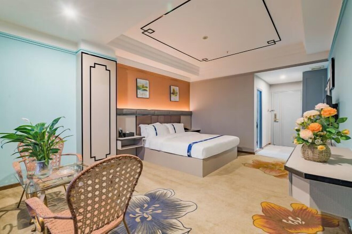 A hotel room with a bed, bedside table, chair, and a small table with a vase of flowers.