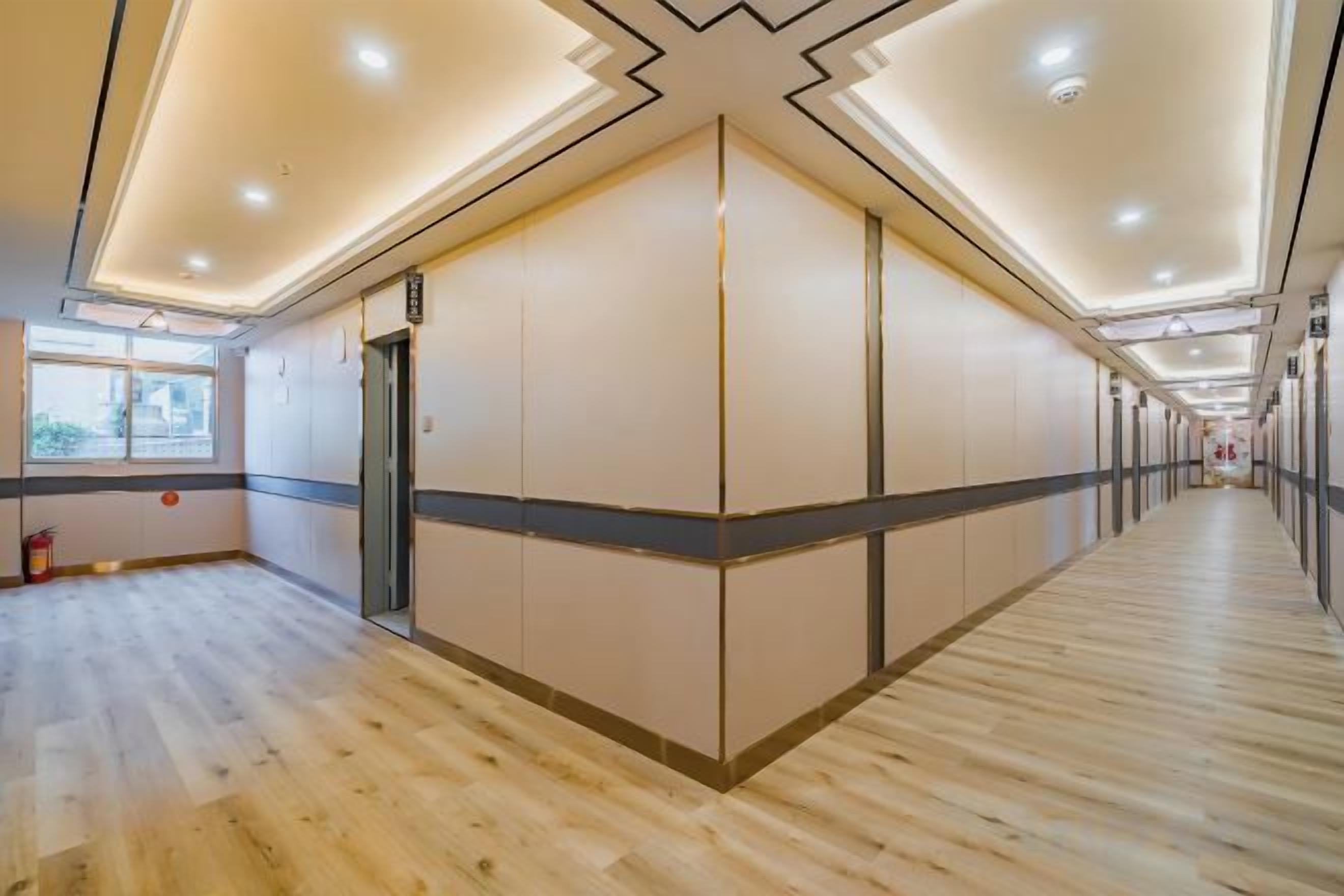 A hotel corridor with wooden flooring, a central elevator, and a long hallway with doors on one side.