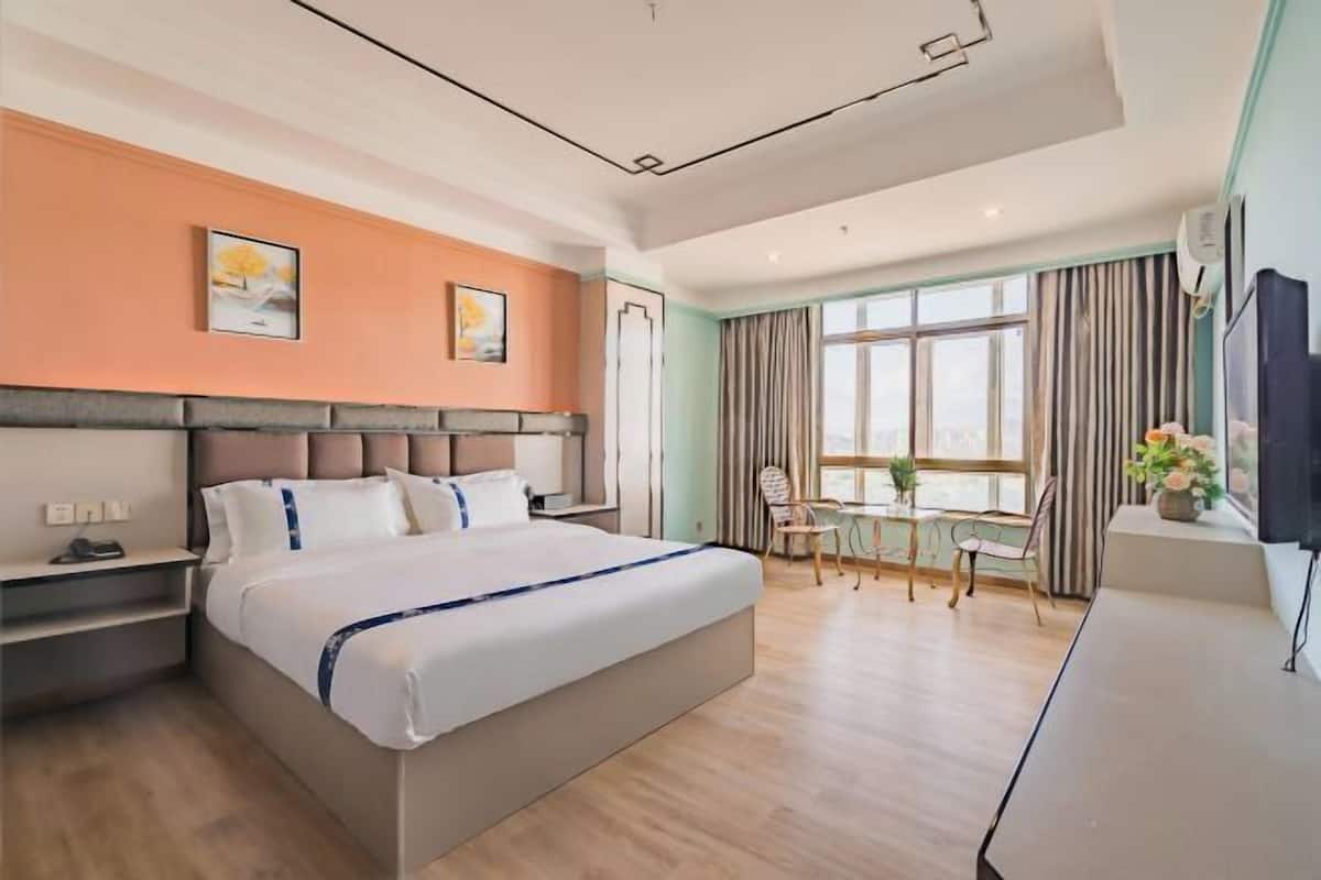 A modern hotel room with a large bed, a desk, and a chair. There is a window with curtains, a TV, and artwork on the wall.