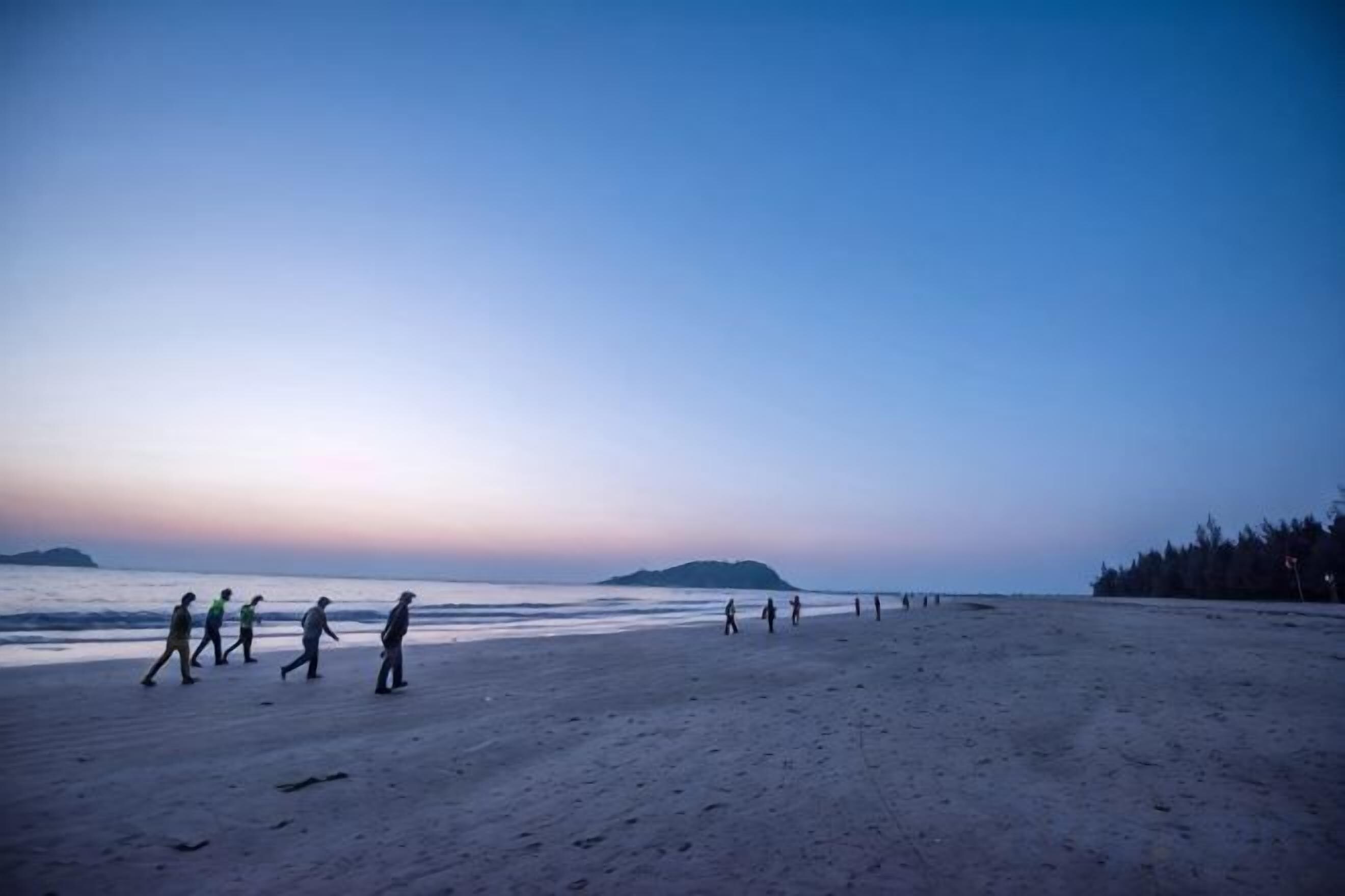 Una playa con gente caminando, cielo despejado y accidentes geográficos a lo lejos.