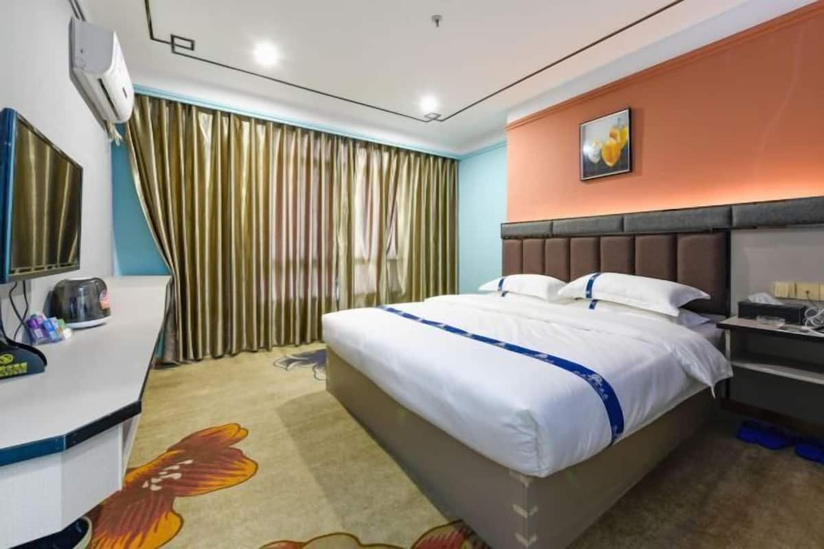 A hotel room with a bed, a desk with a kettle, a television, and a window with curtains.