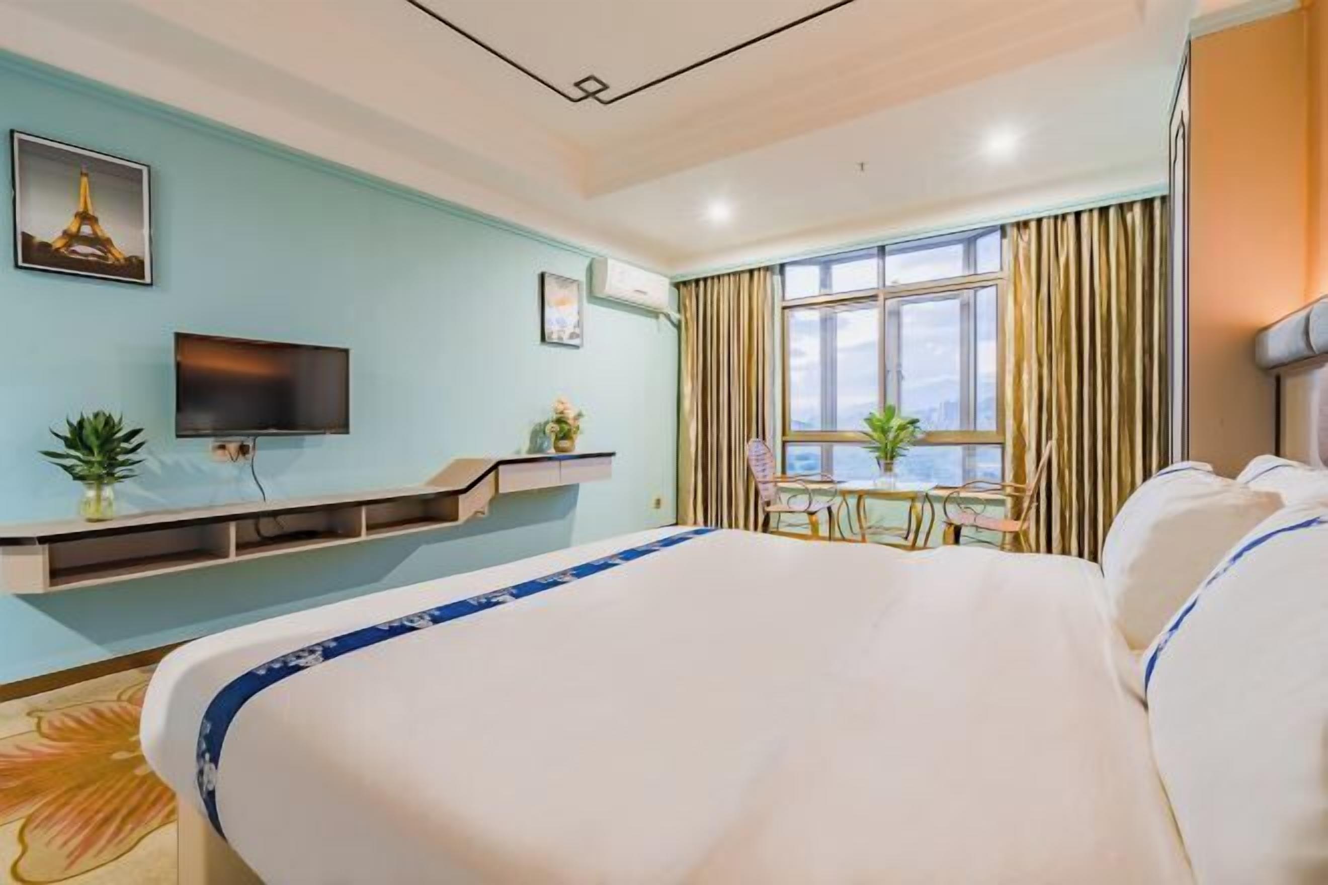 A hotel room with a large bed, a TV mounted on the wall, a desk with a chair, and a window with curtains.