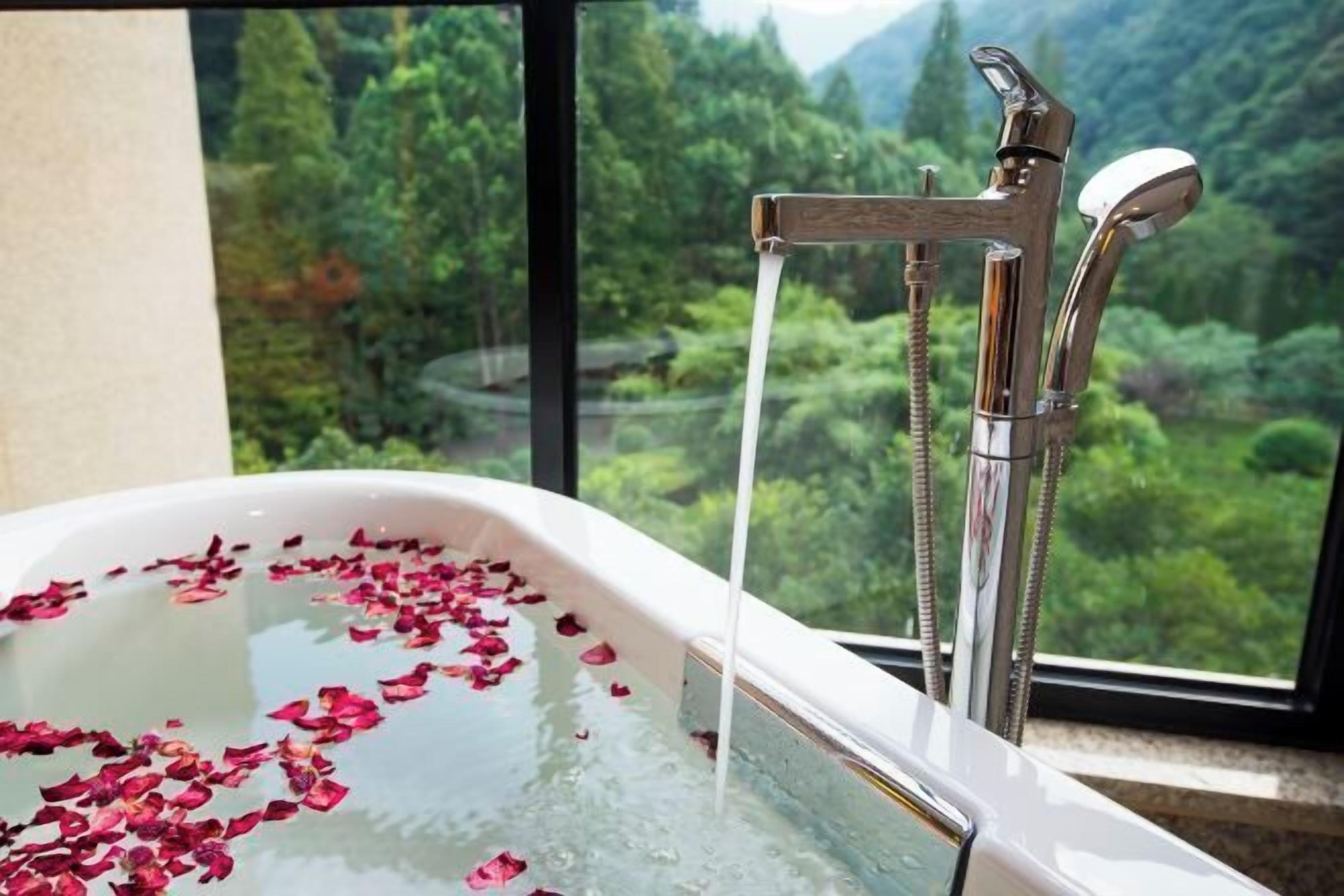 A bathtub with rose petals, a faucet, and a view of a forest through a window.