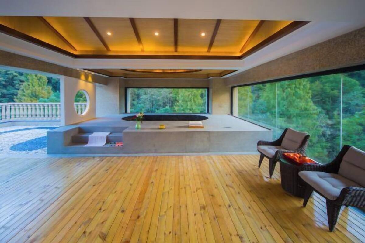 A modern interior with a wooden deck, a seating area with chairs, a built-in bench, and a large window offering a view of greenery.
