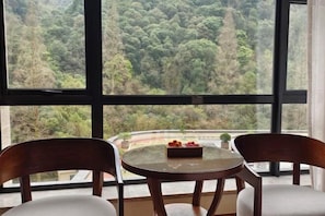 A room with a large window offering a view of a forested area. There is a round table with a tray of fruit and two chairs. - Tianmingshan Spa & Resort (Ninghai)