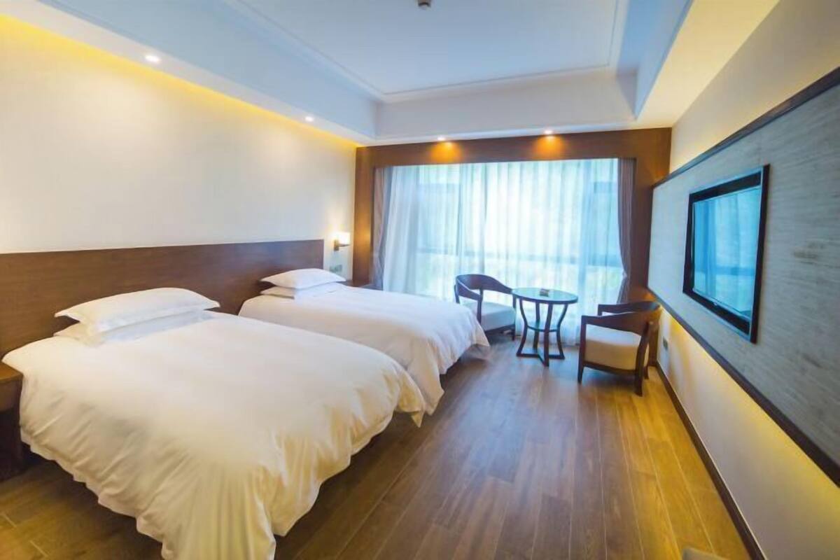 A hotel room with two beds, a small table, a chair, a large window with curtains, and a framed picture on the wall.