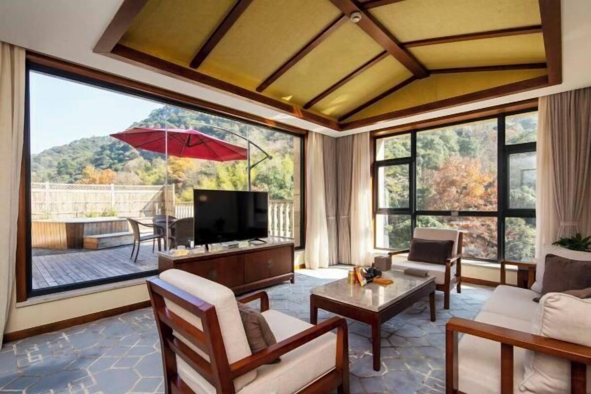 A sunlit room with a TV, wooden furniture, and large windows offering a view of a deck and trees.