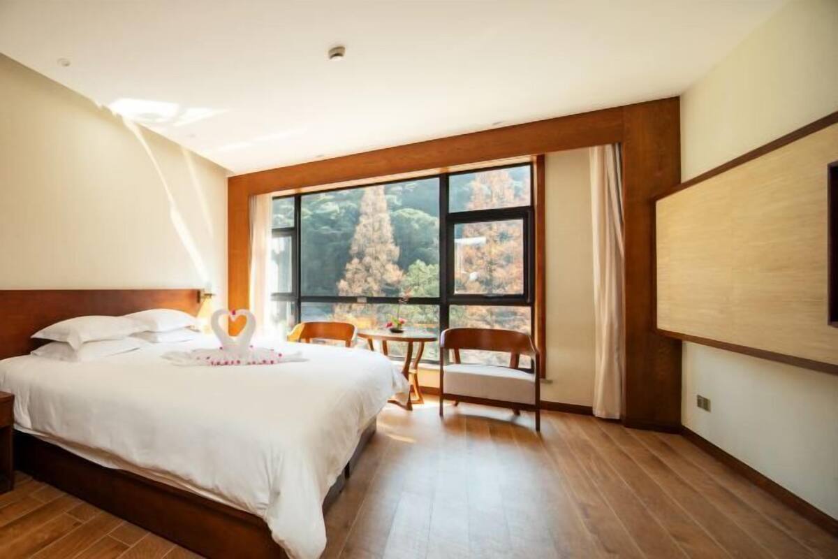 A hotel room with a large bed, a wooden headboard, a small table with chairs, and a large window offering a view of trees.