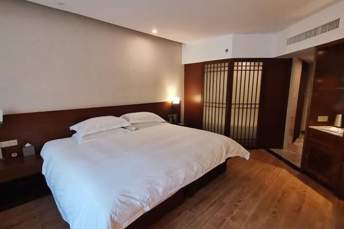 A hotel room with a large bed, wooden flooring, a nightstand, and a wall-mounted lamp.