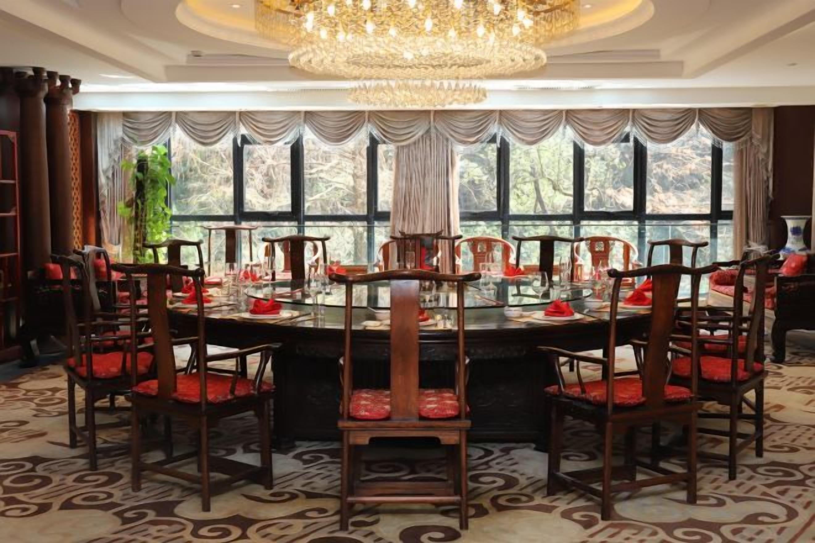 Un comedor con una mesa redonda rodeada de sillas, una lámpara de araña y ventanas grandes con cortinas.