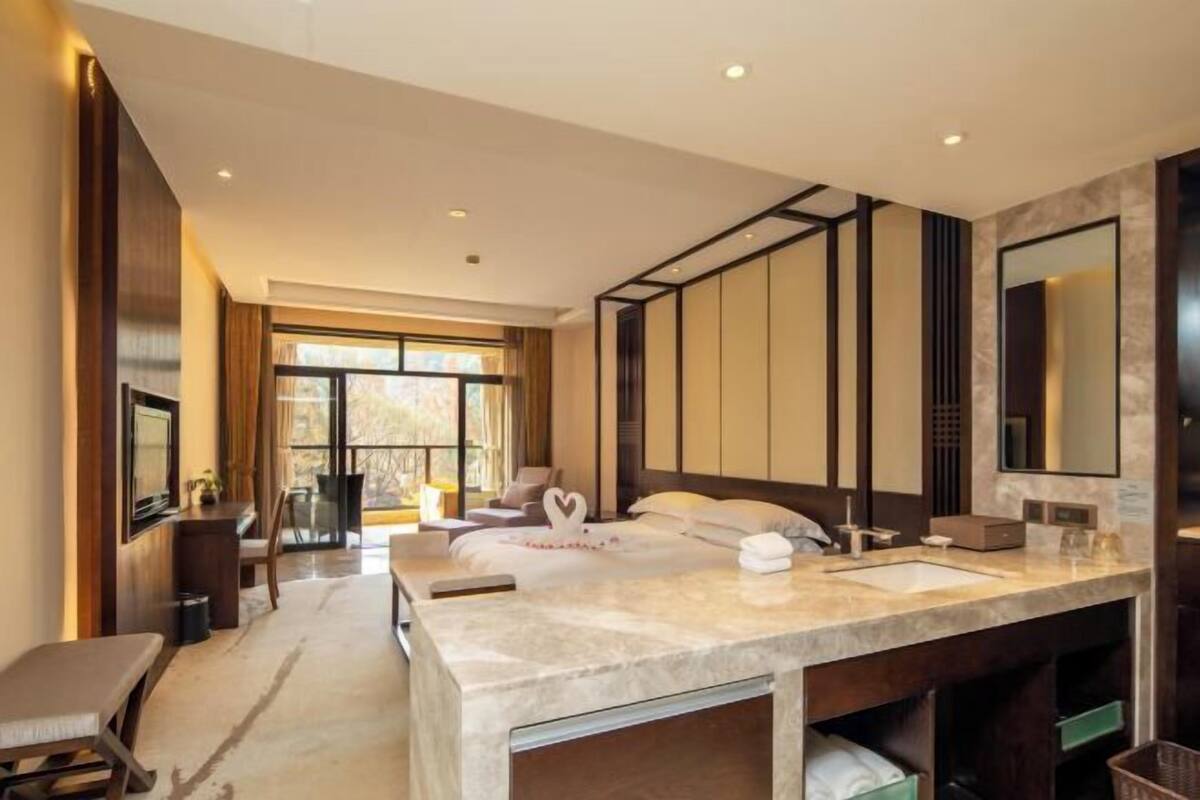A modern hotel room with a large bed, a separate sitting area, a bathroom with a large mirror, and a view of the outdoors.