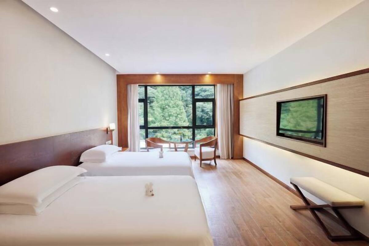 A hotel room with two beds, a TV on the wall, a window with a view of trees, and a small table with chairs.