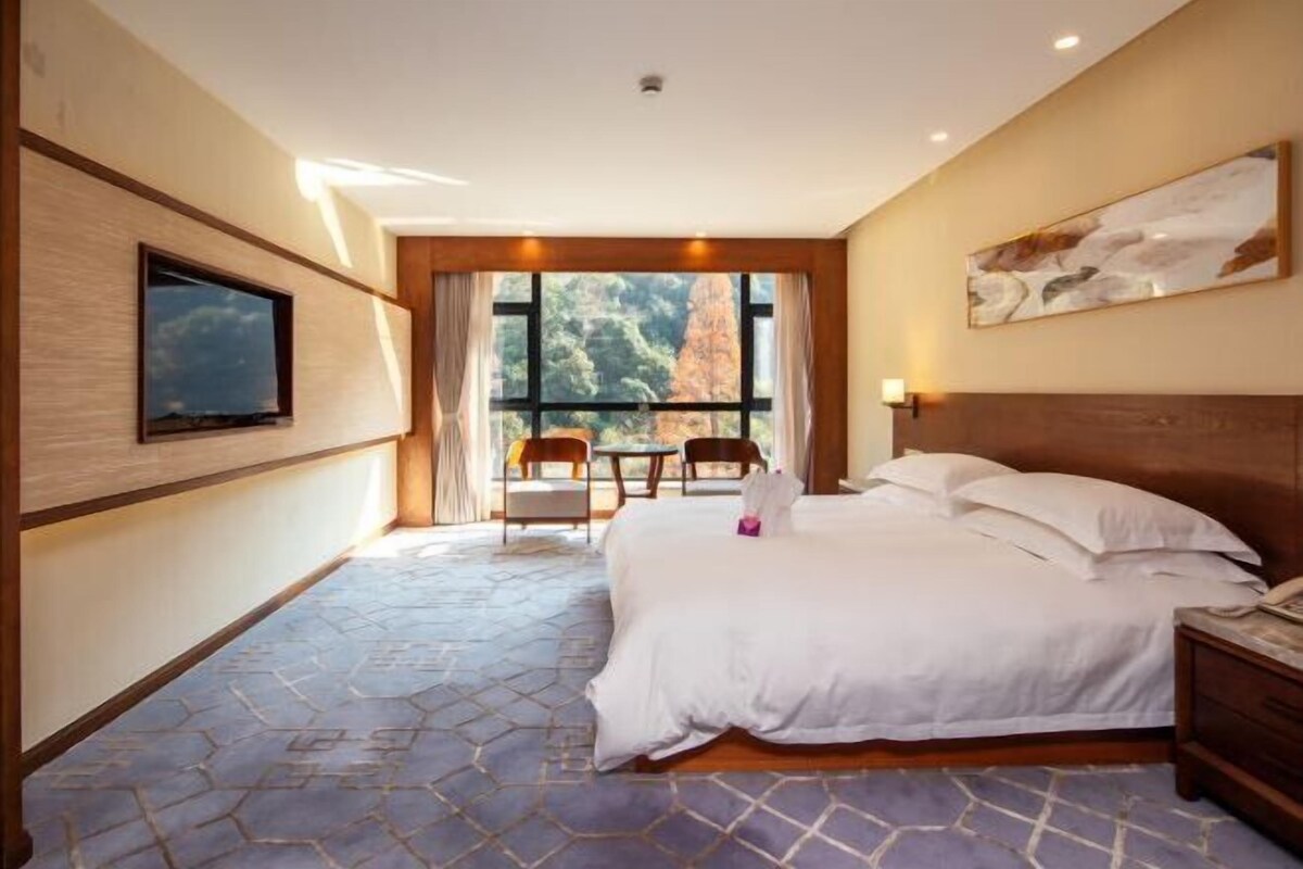 A hotel room with a large bed, a TV mounted on the wall, a window with a view of trees, and a small table with chairs.