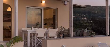 Ein überdachter Sitzbereich im Freien mit Tisch und Stühlen, Blick ins Innere sowie ein Balkon mit Pflanzen und Kissen.