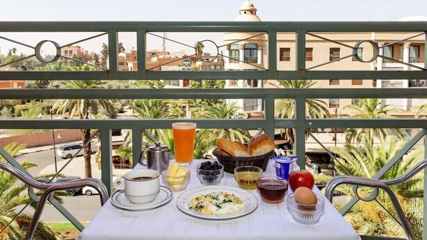 Una mesa en el balcón con alimentos para el desayuno, como huevos, tostadas y bebidas, con vistas a una zona residencial con palmeras y edificios.