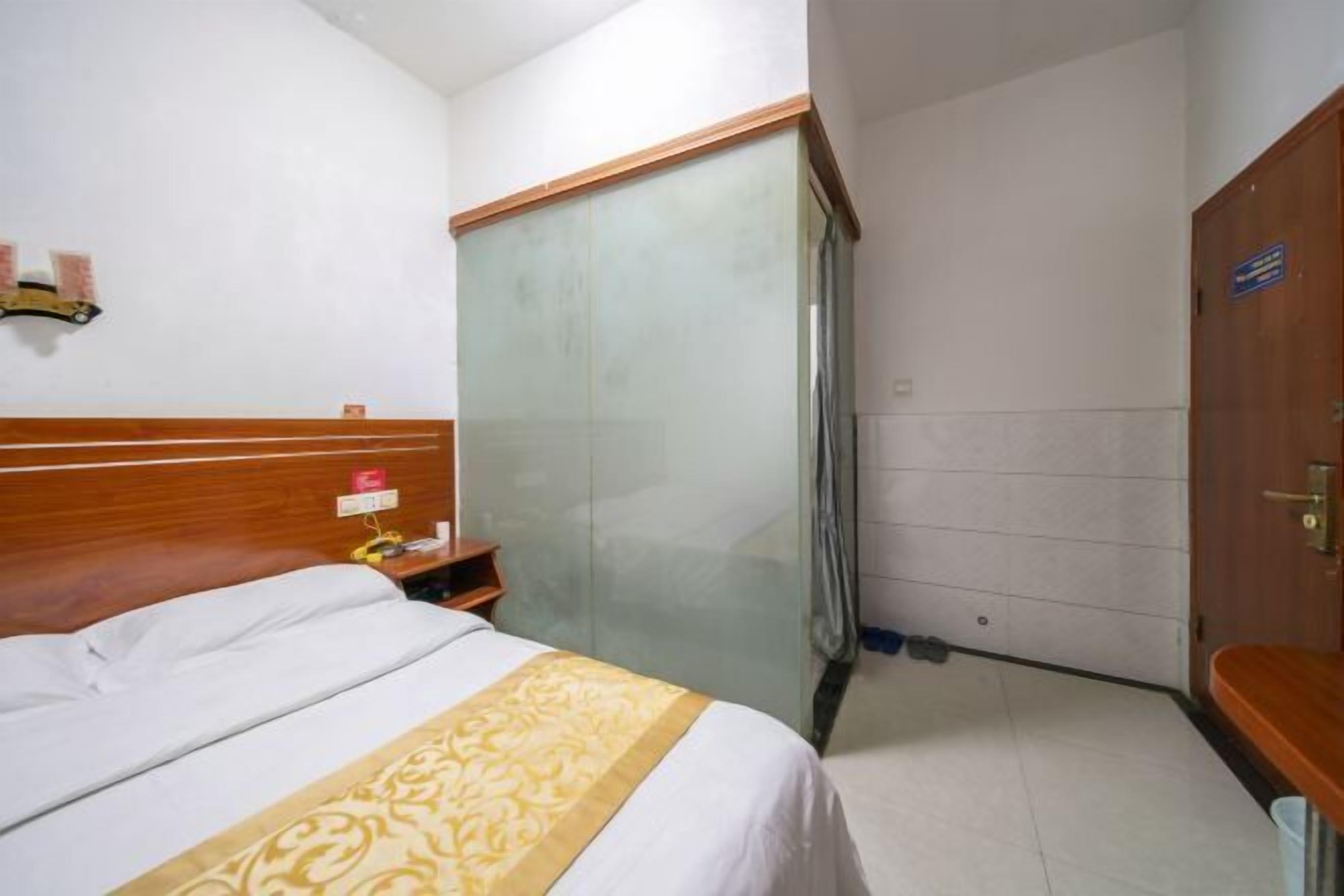 A hotel room with a bed, a wooden headboard, a small table with a lamp, a bathroom with a shower curtain, and a door with a sign.