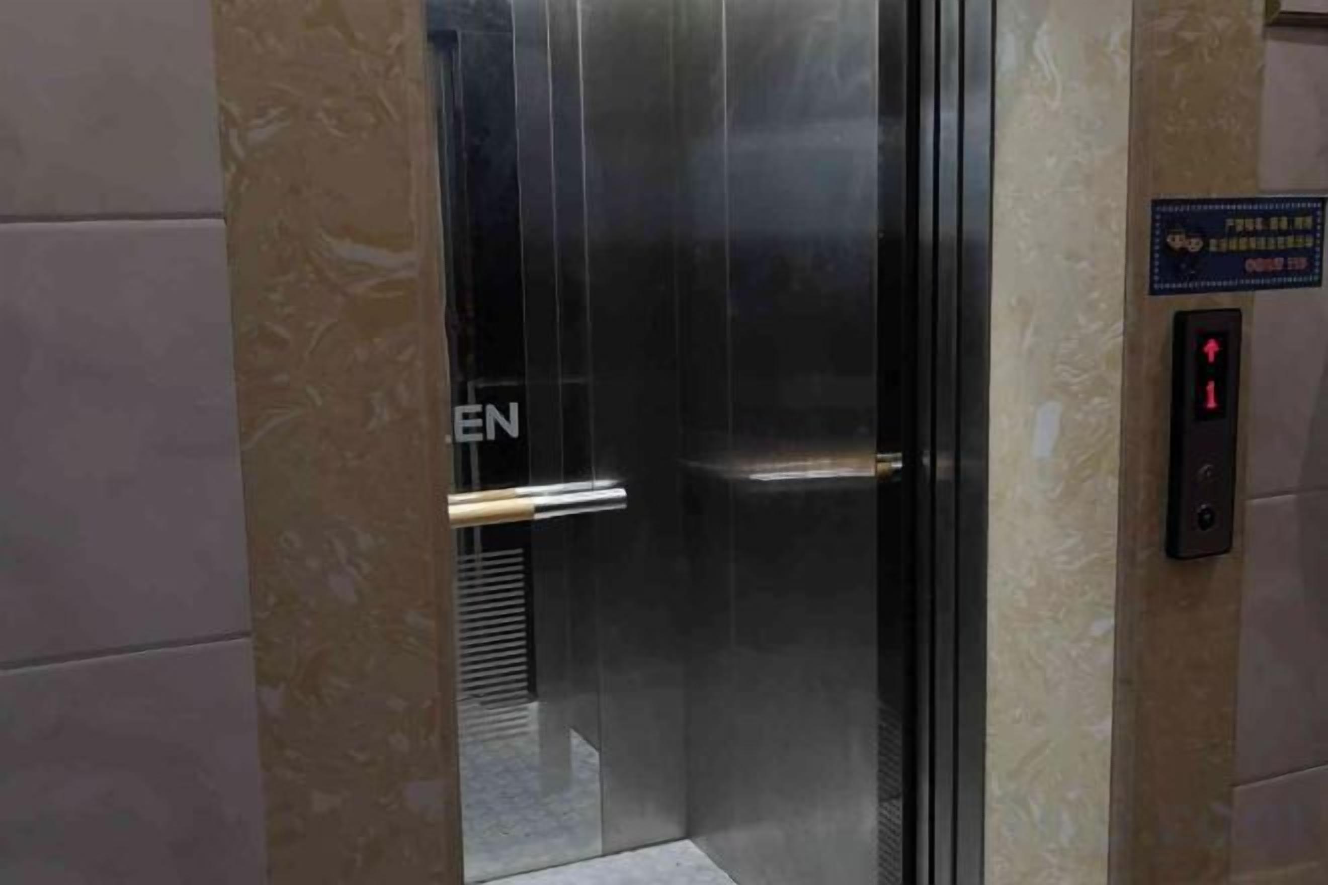 An elevator with a glass door, a visible emergency exit sign, and a control panel with a red light indicator.