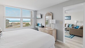 A bedroom with a large bed, a dresser, a mirror, a TV, and a view of the beach. - Sunny 2BR Gulf-View Condo with Balcony & Beach Access | Magnolia House 302 (Destin)
