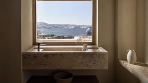 A bathroom with a window overlooking a coastal view, a marble sink, and a vase with dried flowers. - Omeon Mykonos (Mykonos)