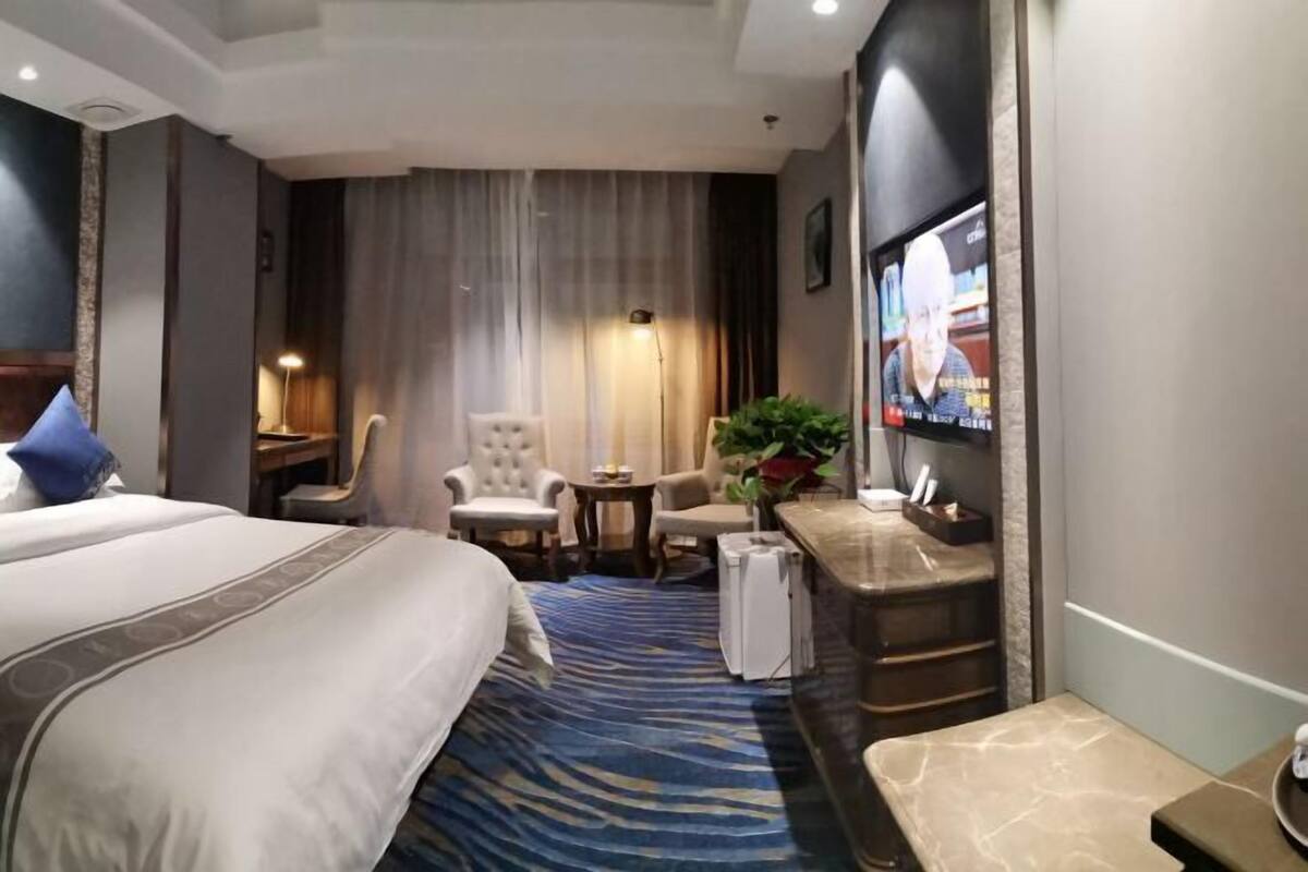 A hotel room with a bed, two armchairs, a small table, a TV, and a plant.