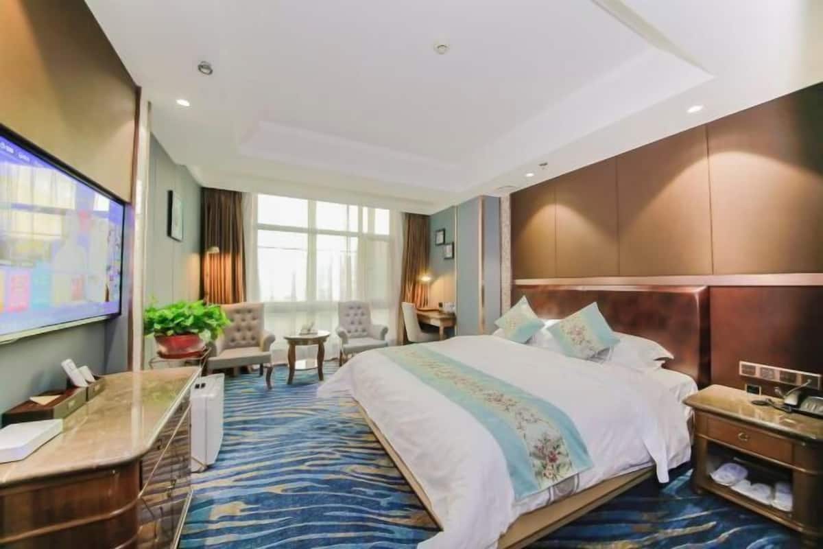 A hotel room with a large bed, a desk with a telephone, a small table with a plant, a TV, and a window with curtains.