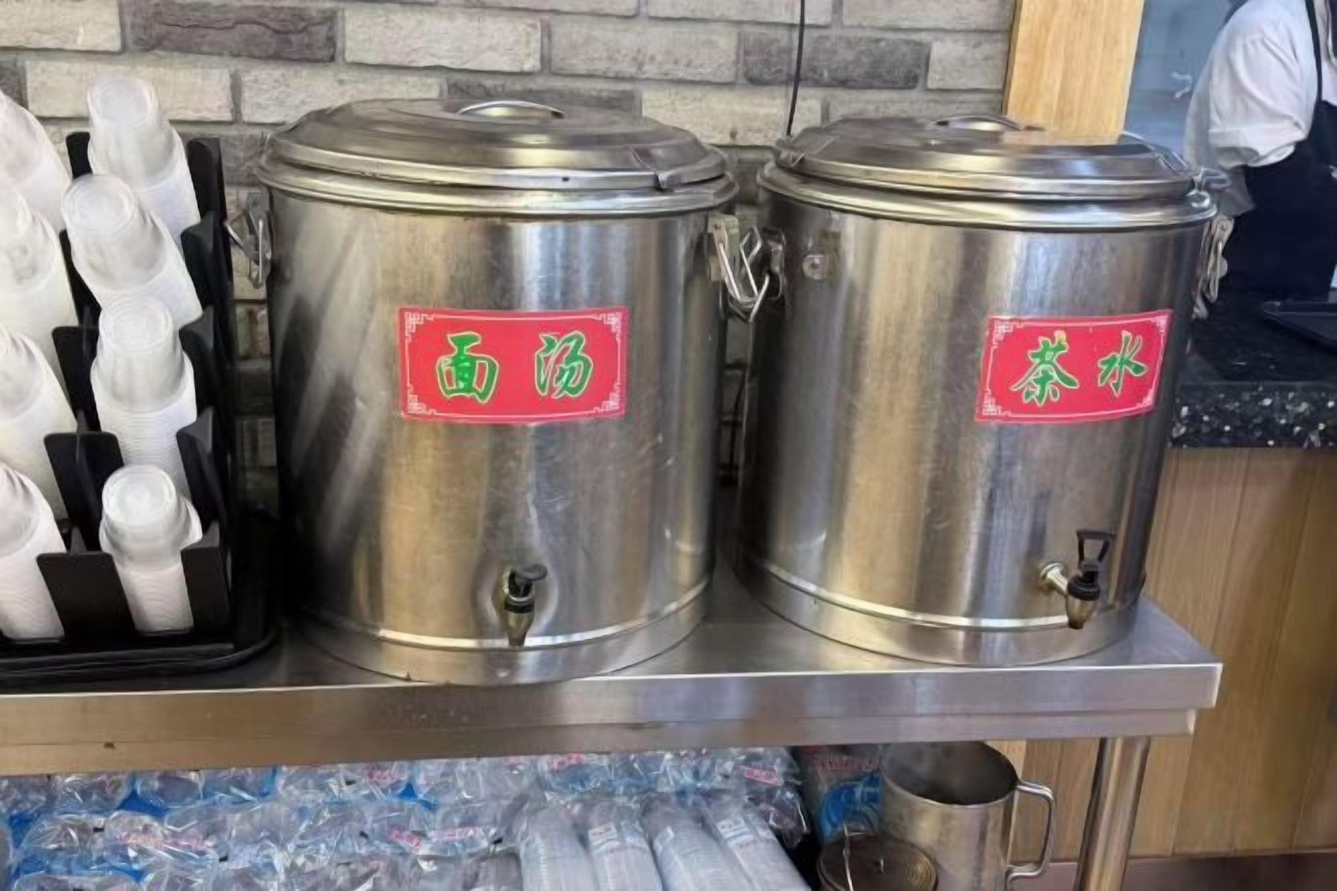 Recipientes de aço inoxidável com etiquetas, uma bandeja de copos descartáveis brancos e uma bancada.
