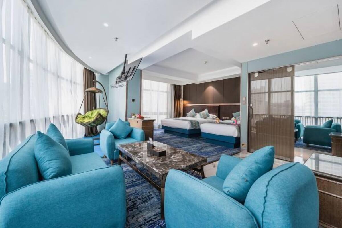 A modern hotel room with a large bed, a seating area with blue sofas, a coffee table, and a balcony with a view.
