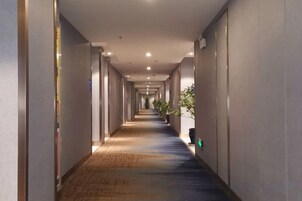 Un largo y estrecho pasillo de hotel con piso alfombrado, paredes con puertas y plantas en macetas a la derecha.