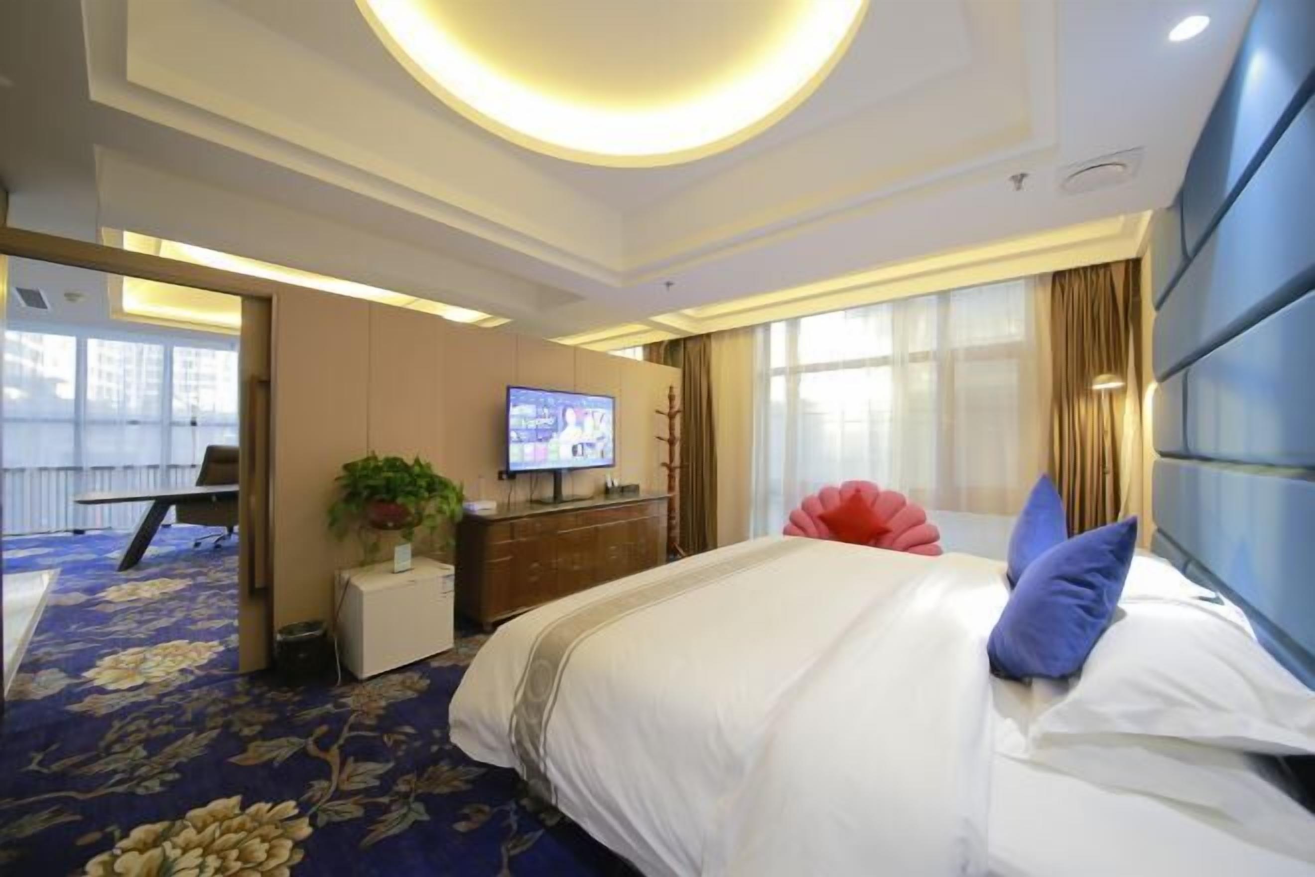 A hotel room with a large bed, a flat-screen TV, a desk, and a potted plant.