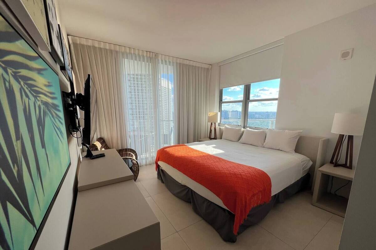 A hotel room with a large bed, a desk, a chair, a TV, and a window with a city view.