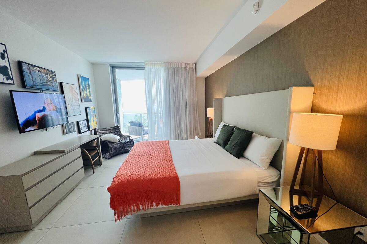 A modern hotel room with a large bed, a TV mounted on the wall, a desk with a chair, and a balcony with a view.