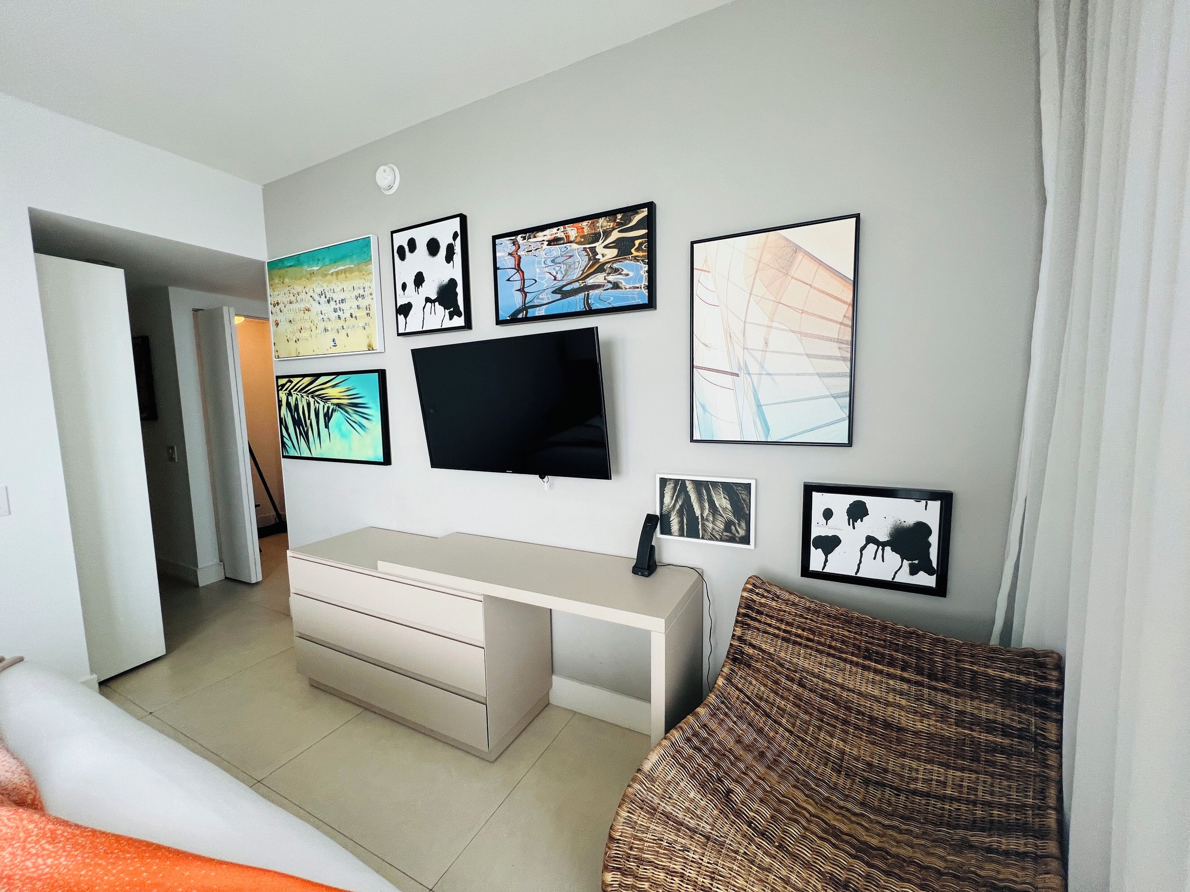 Une chambre moderne avec un grand lit, un bureau blanc et un téléviseur. Plusieurs tableaux encadrés ornent le mur.